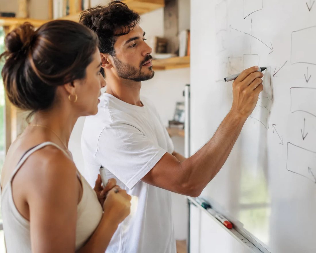Duas pessoas em pé diante de um quadro branco onde um argumento está desenhado